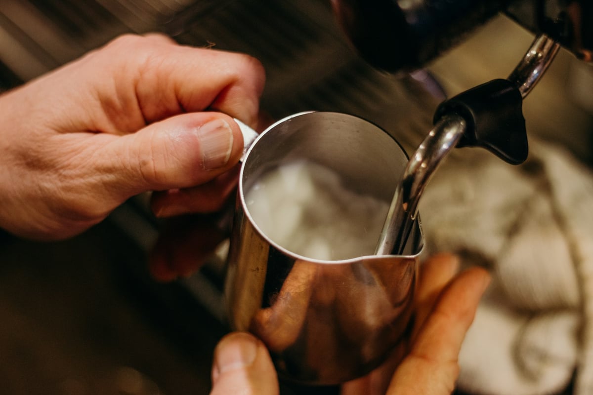 Hands holding a stainless steel pitcher while a steam wand textures milk for espresso