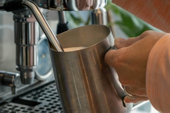 Hand steaming milk in a stainless steel pitcher under a home espresso machine steam wand