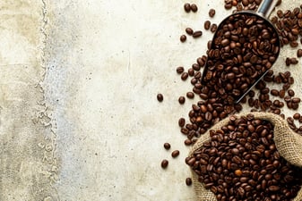Metal scoop of dark roasted coffee beans beside a burlap sack of coffee beans