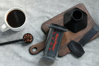 AeroPress coffee maker with funnel, stirrer and scoop of beans beside a cup of black coffee