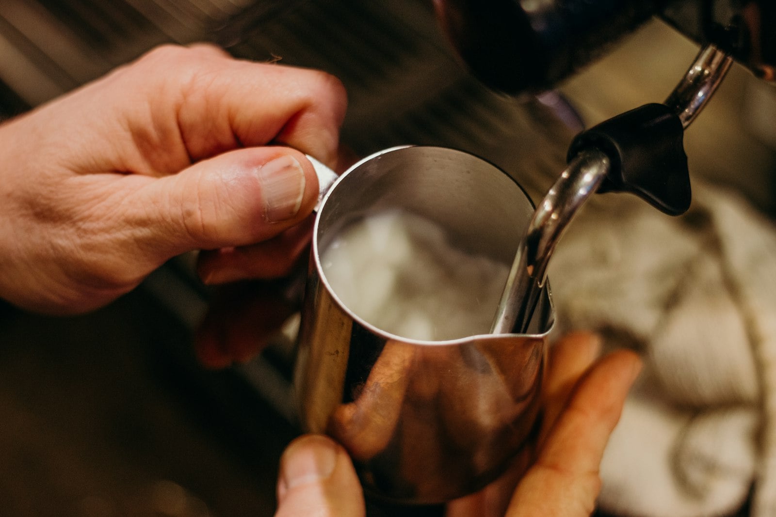 Hands holding a stainless steel pitcher while a steam wand textures milk for espresso